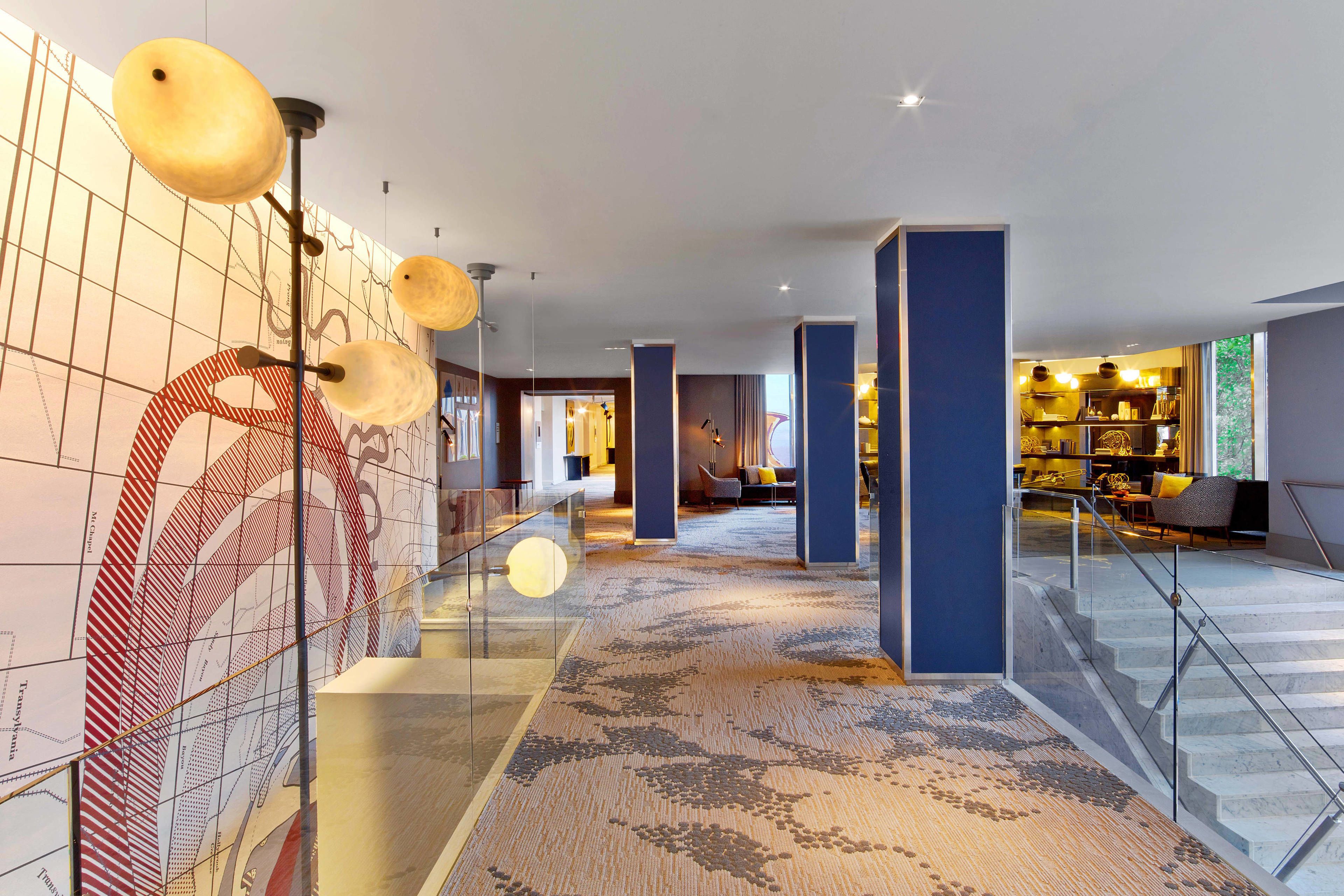 A modern hotel lobby with a patterned carpet, glass railings, and a staircase leading to an upper level.