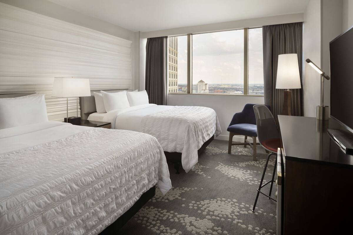 Ein Hotelzimmer mit zwei Betten, einem Schreibtisch, einem Sessel und einem großen Fenster mit Blick auf die Stadt.