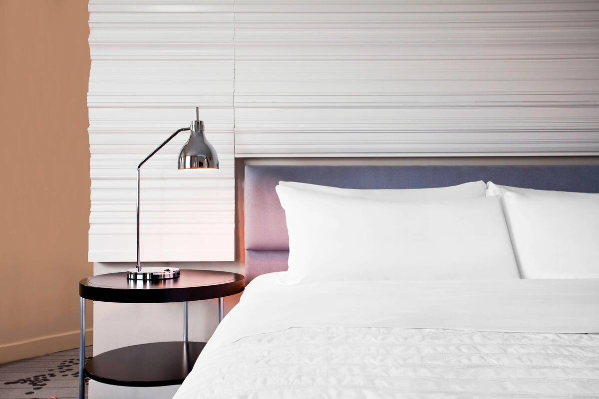 Ein ordentlich bezogenes Bett mit weißen Laken, ein runder Nachttisch mit Lampe und ein Fenster mit Jalousien.