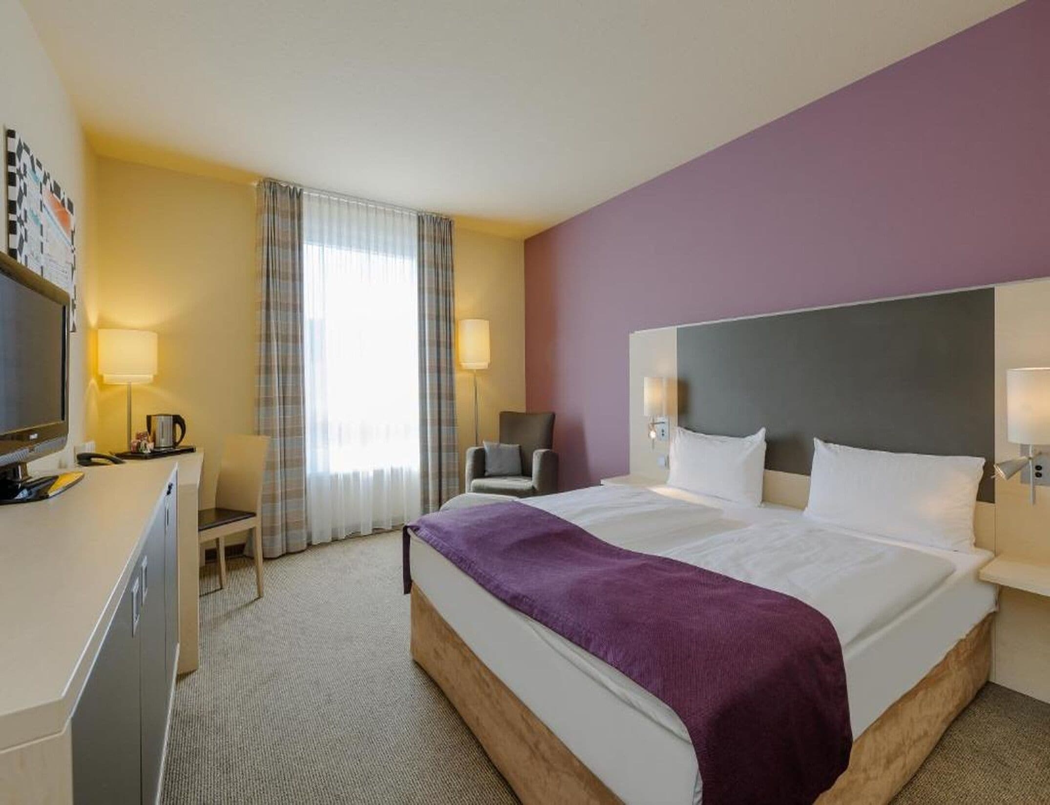 Ein Hotelzimmer mit Bett, Schreibtisch, Sessel, Fernseher und einem Fenster mit Vorhängen.