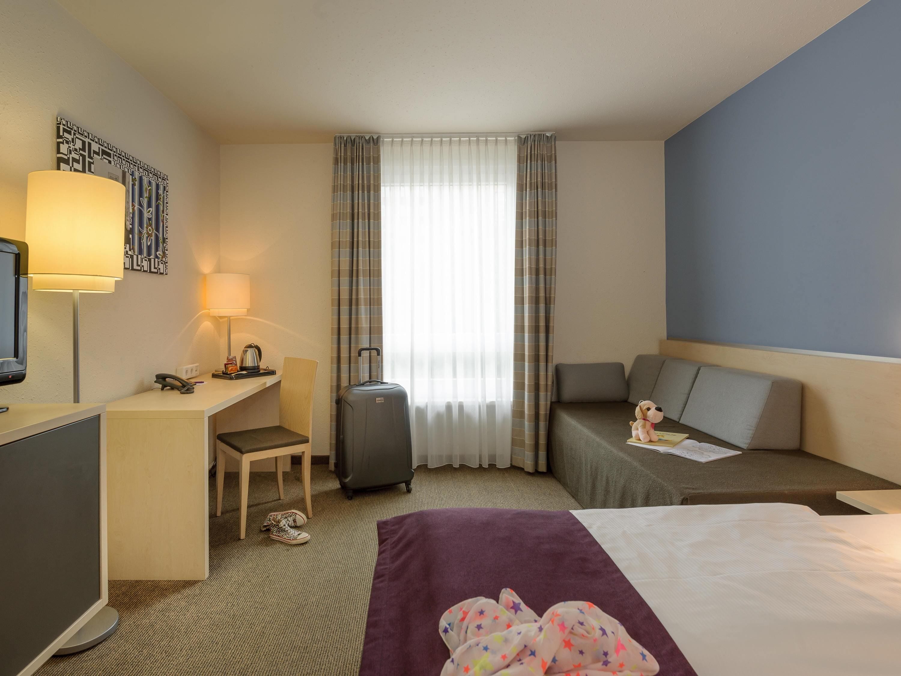A hotel room with a desk, chair, bed, and a suitcase.