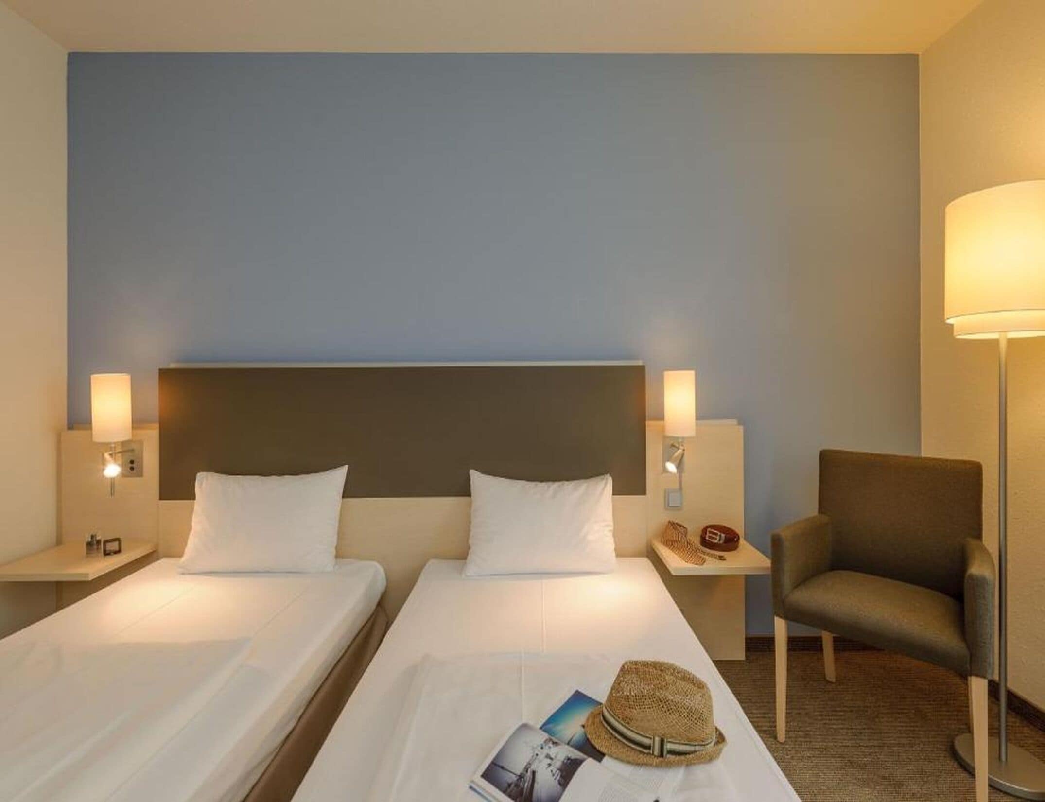 Una habitación de hotel con dos camas, una mesita de noche con una lámpara, una silla y un sombrero sobre la cama.