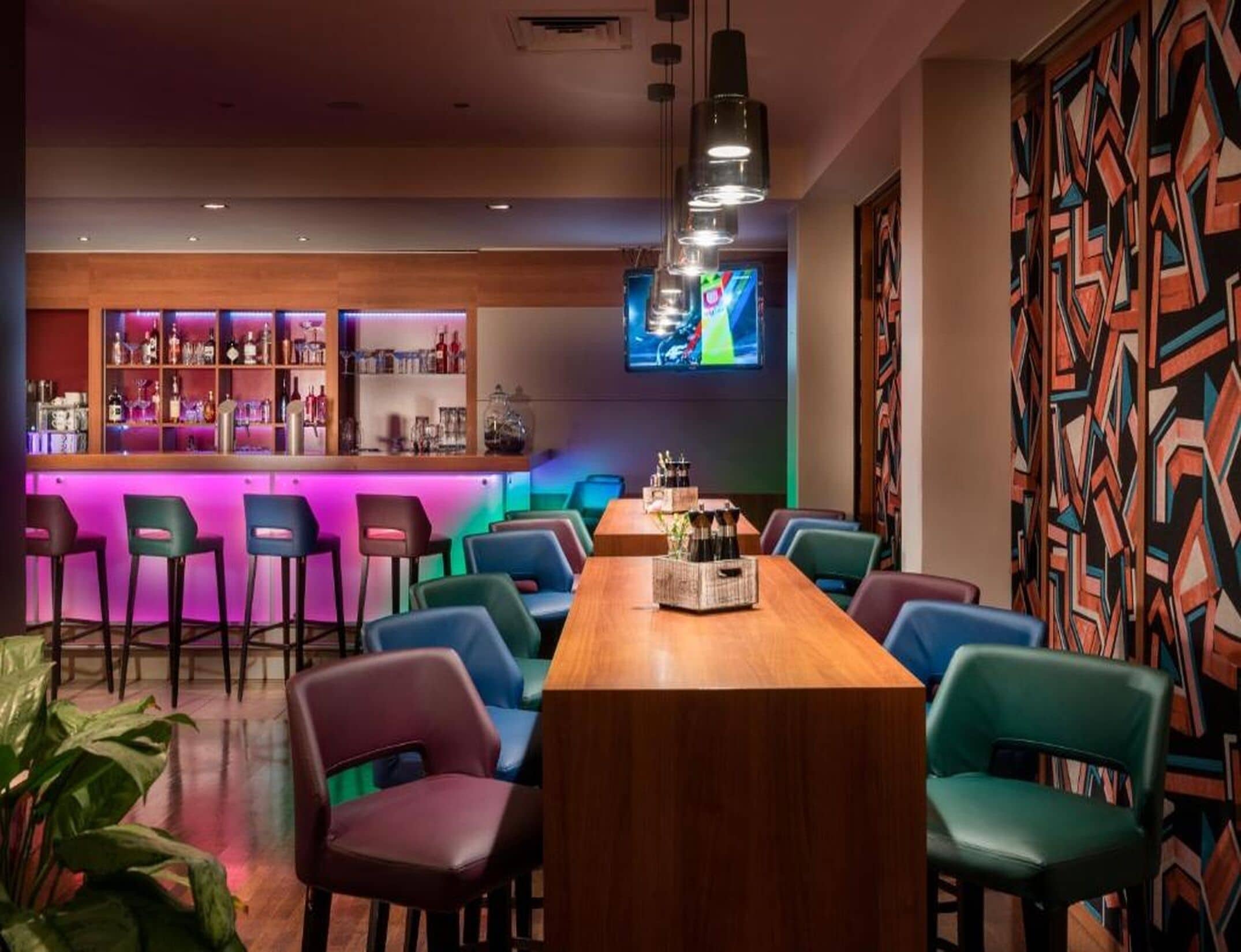 A bar area with a long wooden table, colorful chairs, and a bar stocked with bottles.