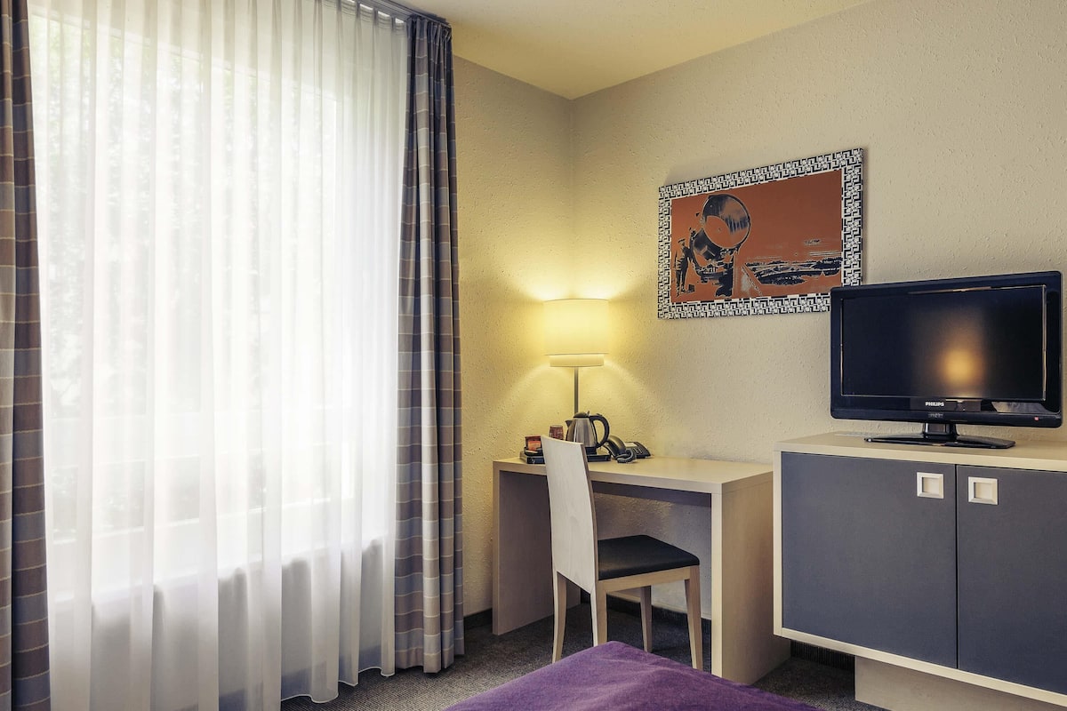 Une chambre d’hôtel comprenant un lit, un bureau avec une chaise, une télévision, une lampe et une fenêtre avec des rideaux.