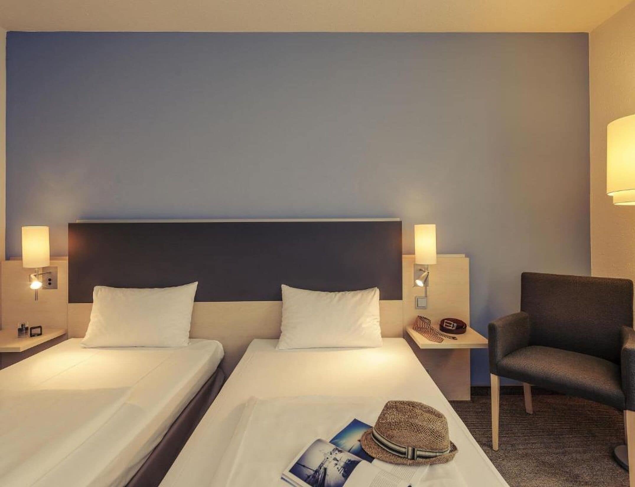 Habitación de hotel con dos camas, una mesita de noche con lámpara, una silla y un sombrero sobre la cama.