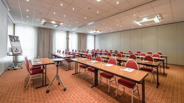 A conference room with multiple tables and chairs, a projector screen, and a carpeted floor. - Sunday Hotel Berlin City West (Berlin)