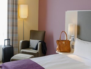 Una habitación de hotel con una cama, una silla, una maleta, un bolso y una lámpara.