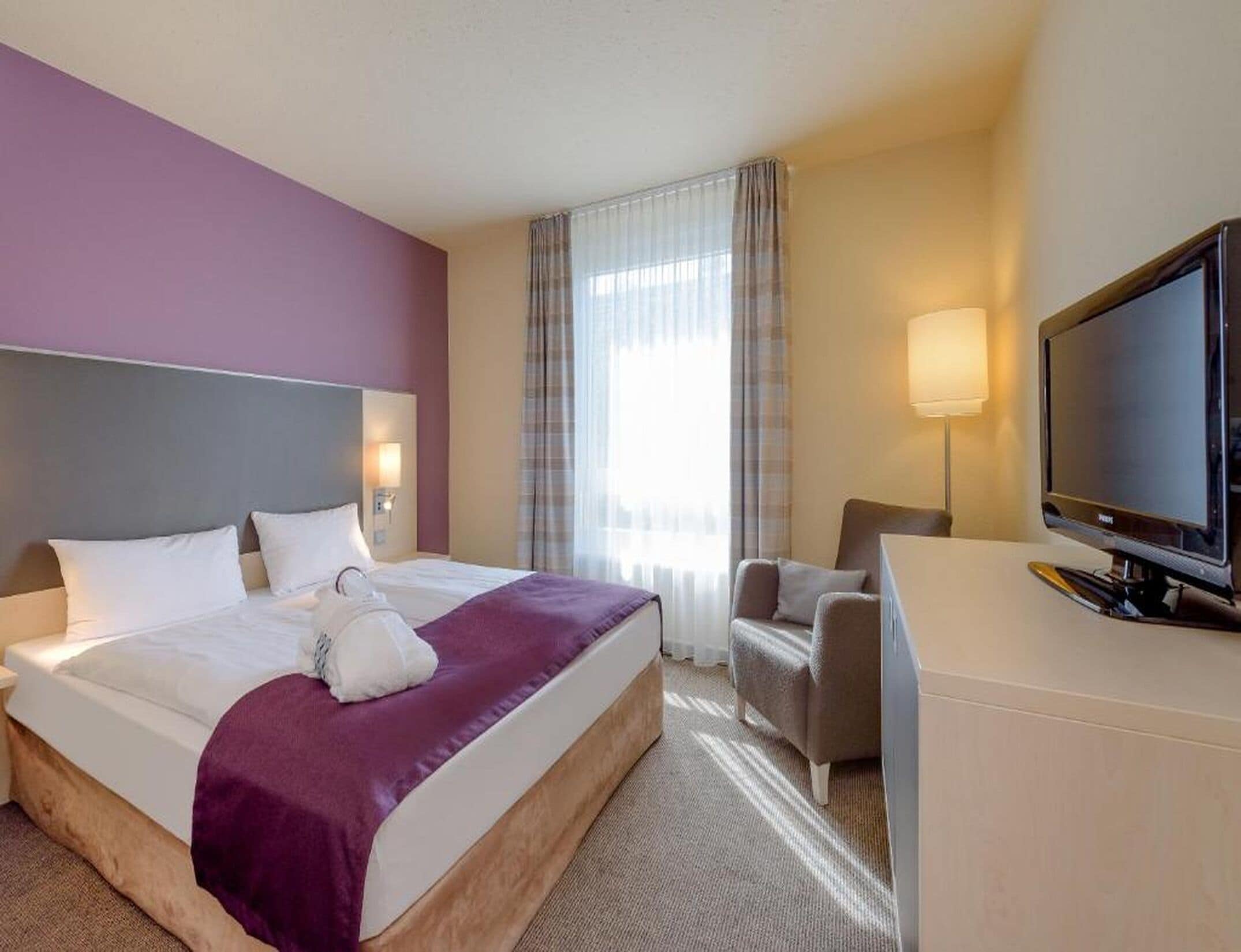 Une chambre d’hôtel avec un lit, un bureau, une chaise, une télévision et une fenêtre avec des rideaux.