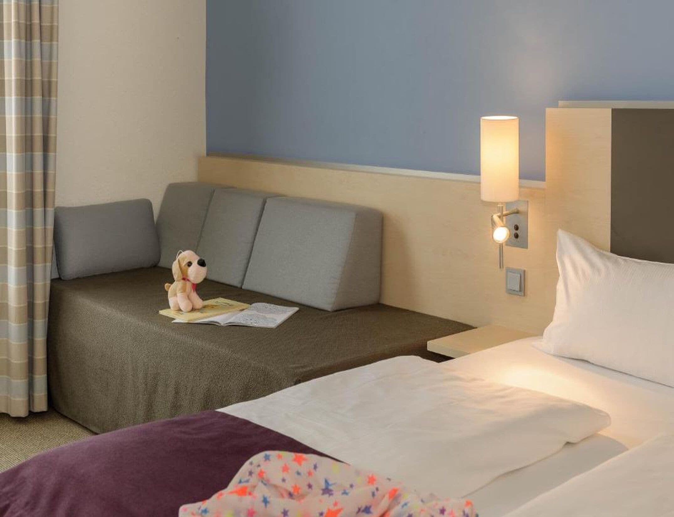 Habitación de hotel con cama, sofá, mesita de noche, lámpara y un pequeño peluche.