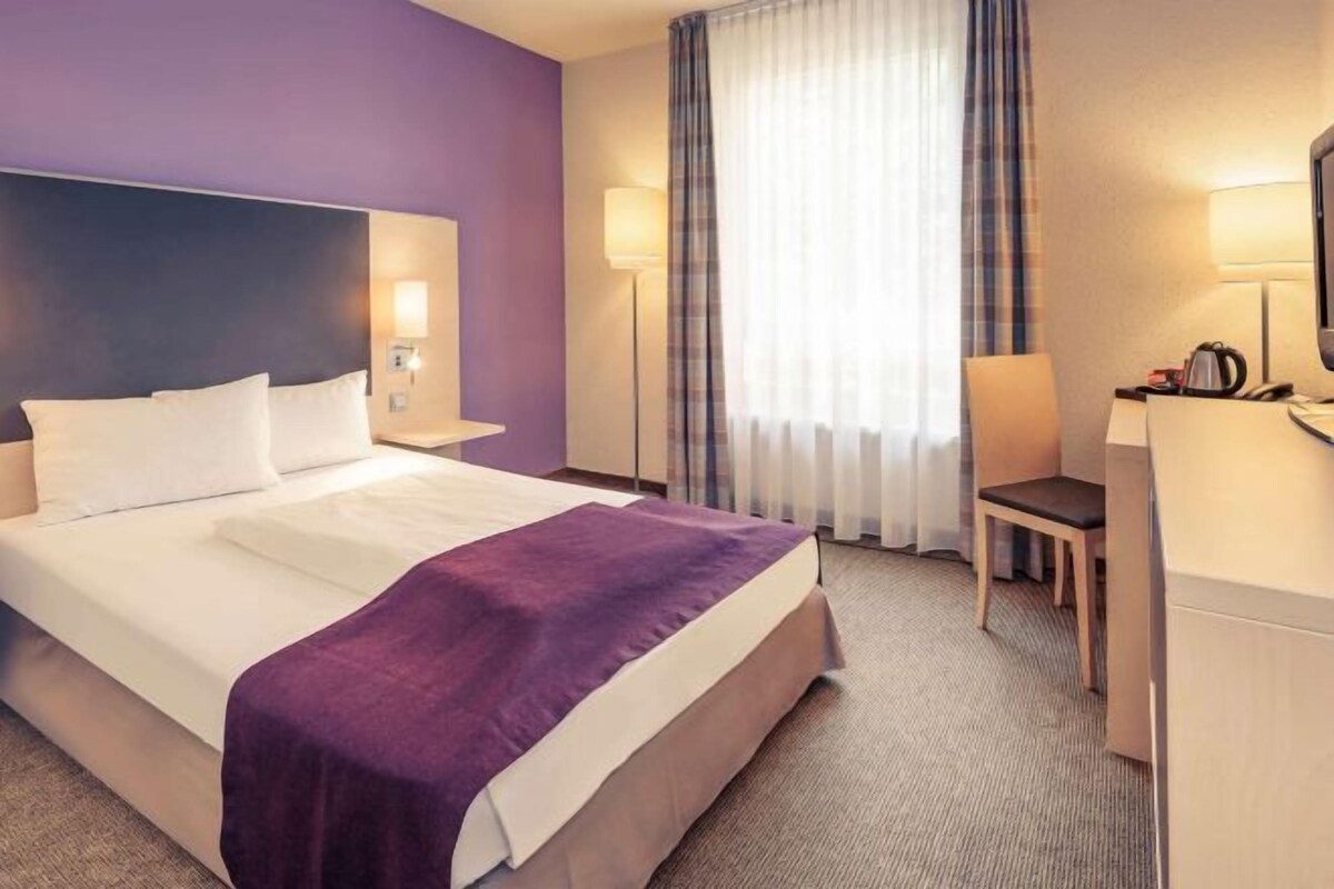 Una habitación de hotel con cama, escritorio, silla y ventana con cortinas.