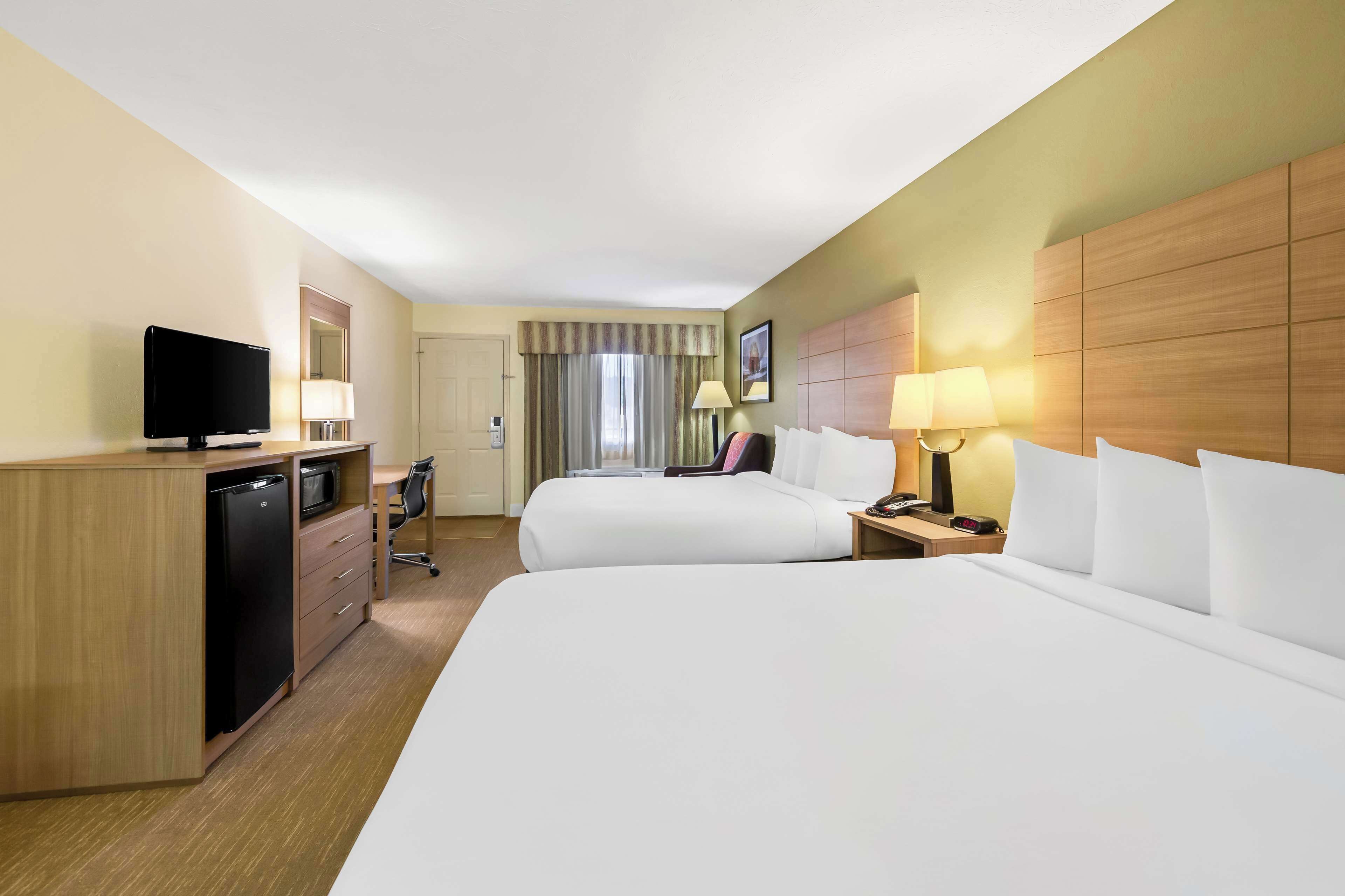 Habitación de hotel con dos camas, un escritorio con televisor, un mini refrigerador, una lámpara y una mesita de noche.