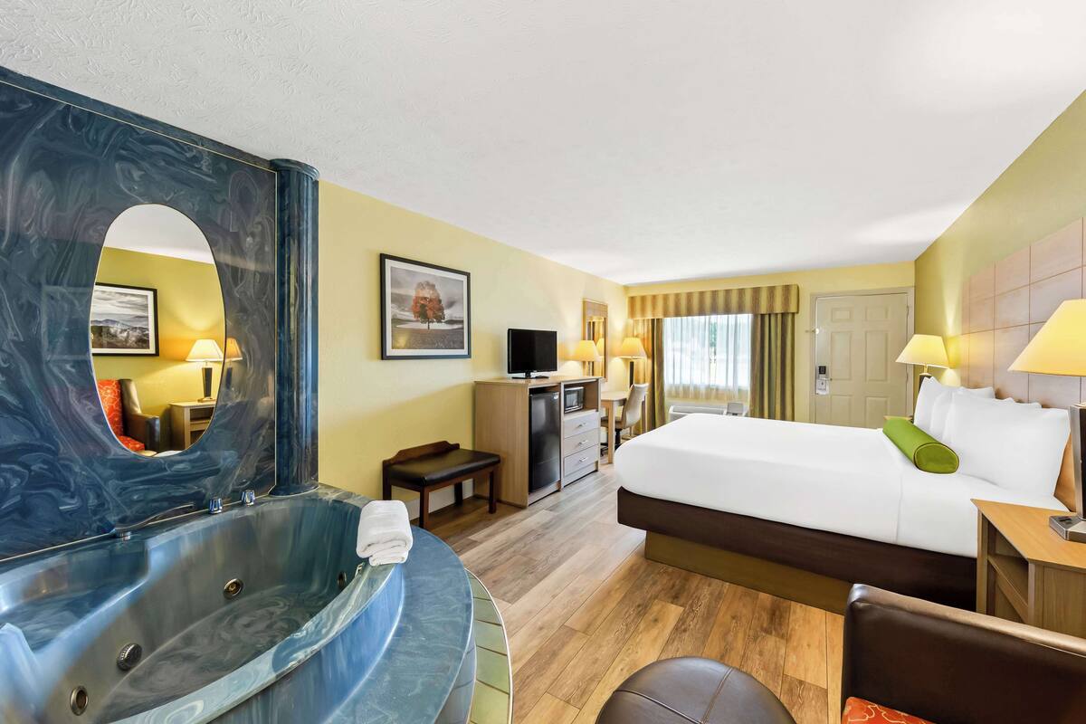 A hotel room with a large bed, a sitting area with a desk and chair, a television, a minibar, and a large mirror.