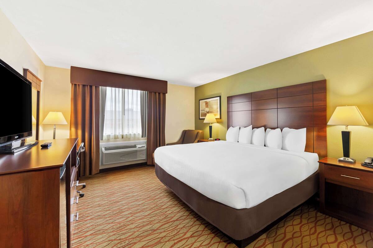 A hotel room with a large bed, a desk with a TV, a chair, a bedside table with a lamp, and an air conditioning unit.