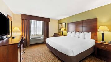 A hotel room with a large bed, a desk with a TV, a chair, a bedside table with a lamp, and an air conditioning unit.
