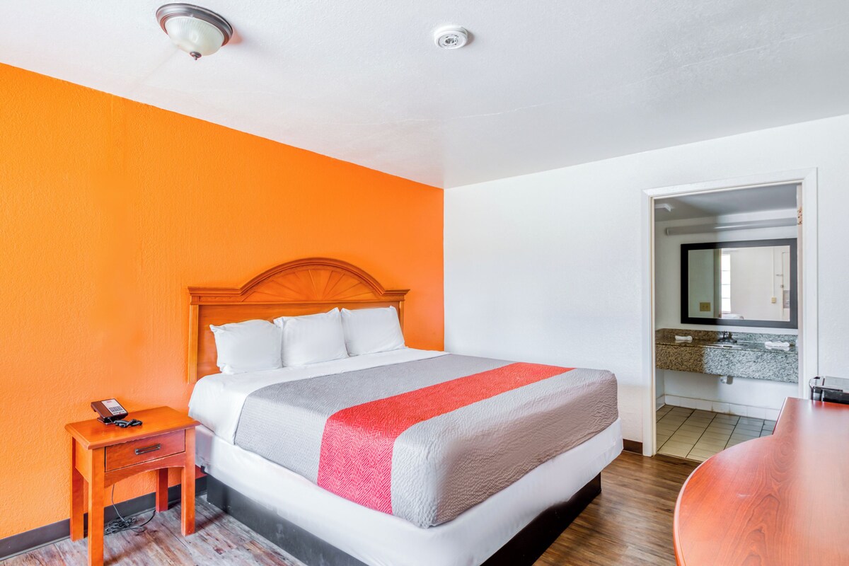A hotel room with a bed, a bedside table with a phone, a wooden nightstand, a bathroom with a sink and mirror, and a wooden floor.