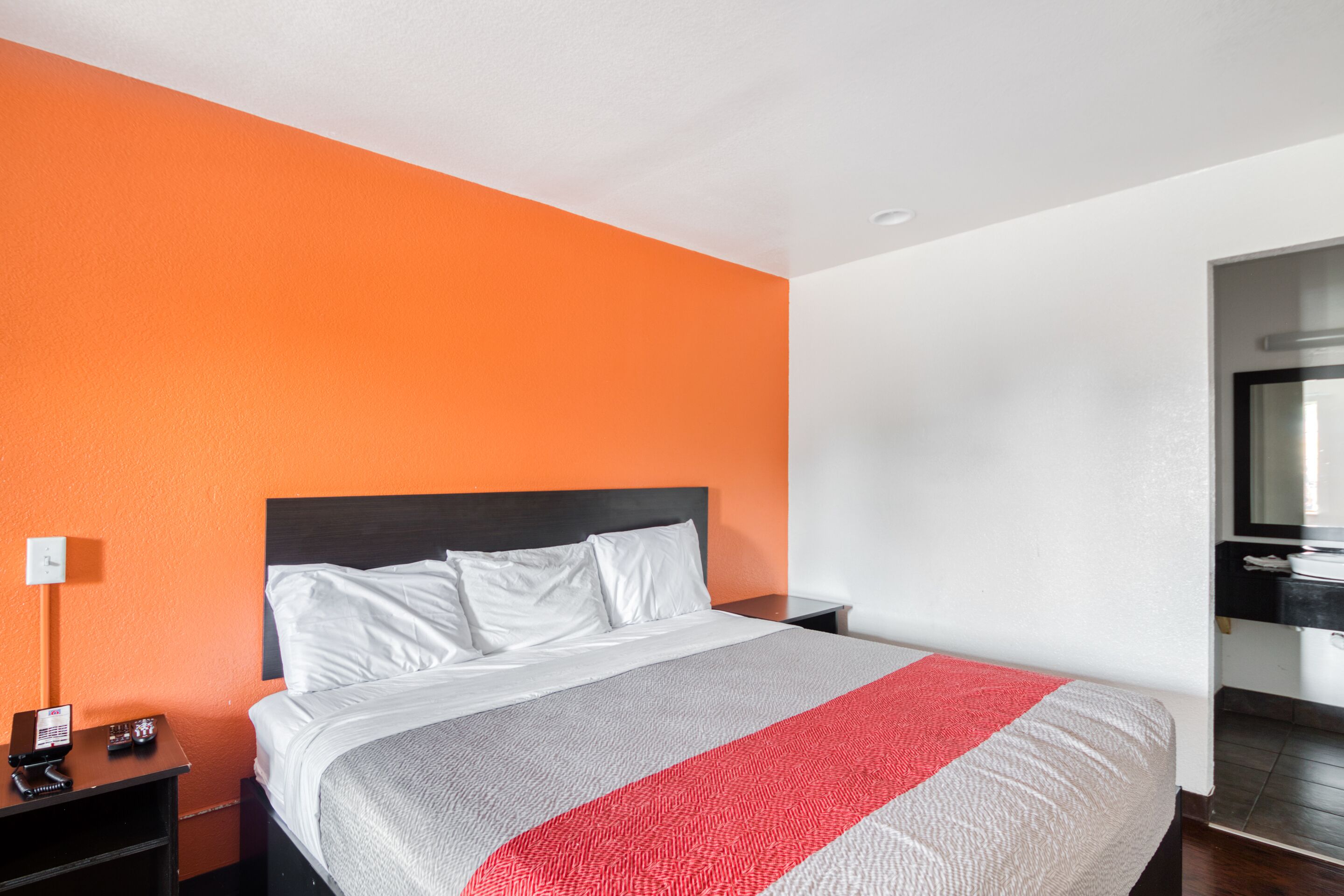 A hotel room with an orange accent wall, a large bed with white bedding, a nightstand with a phone and a lamp, and a bathroom with a mirror and sink.