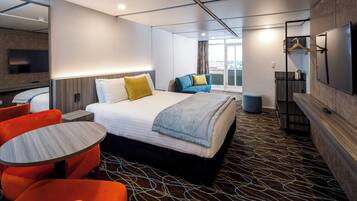 A hotel room with a large bed, a seating area with orange chairs, a small table, a TV, and a view of the city through the window.