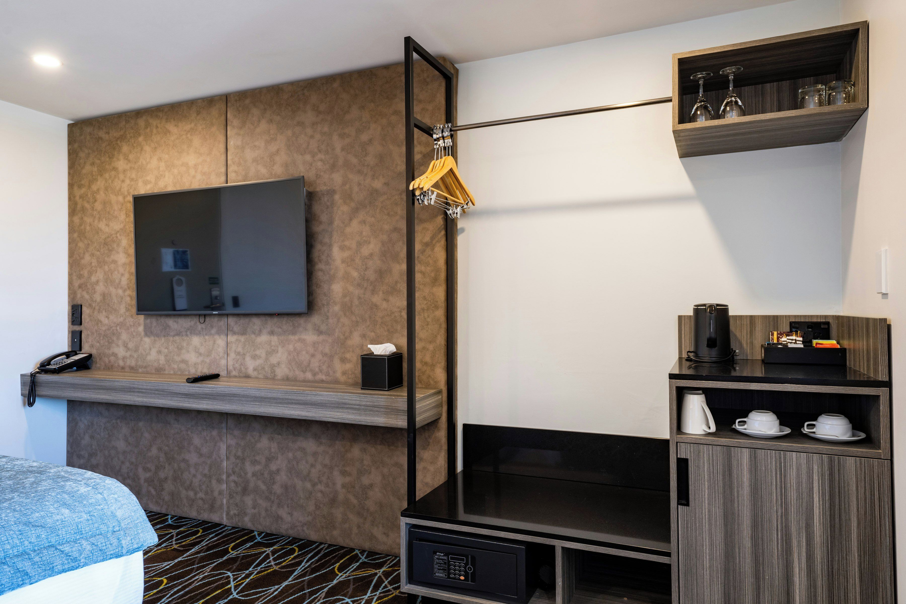 A modern hotel room with a flat-screen TV mounted on the wall, a wooden shelf with a telephone and remote control, a black cabinet with a coffee maker and cups, and a patterned carpet.