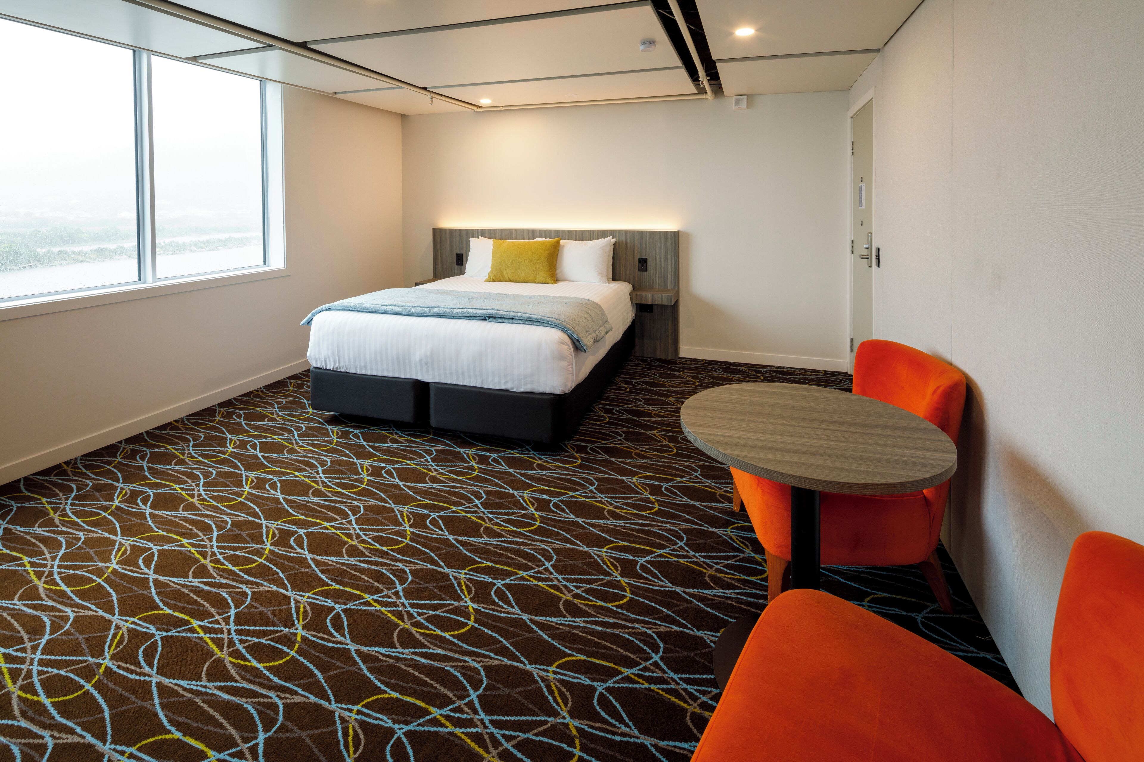 A hotel room with a large bed, a bedside table, a chair, and a view of the outdoors through a window.