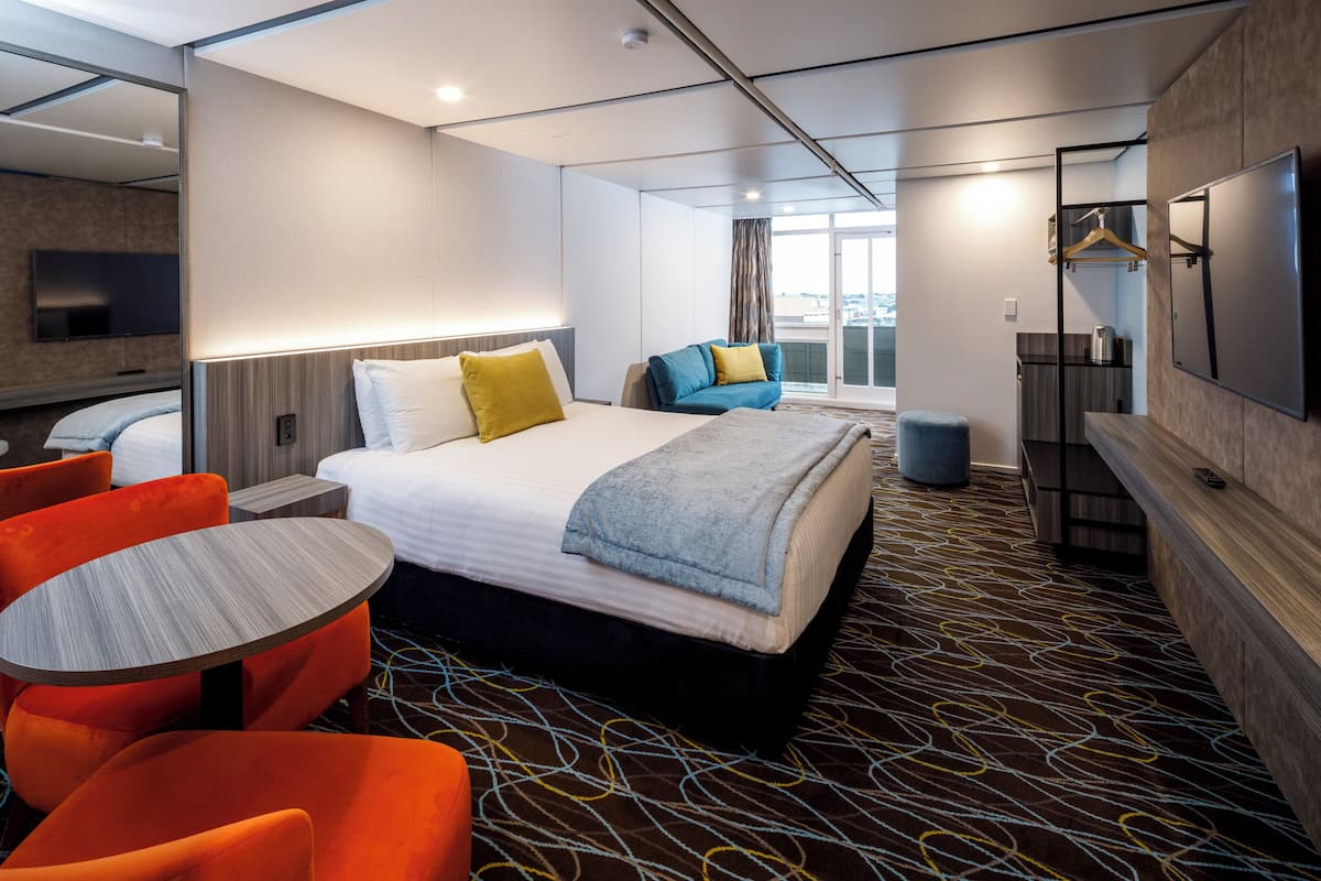 A hotel room with a bed, a seating area with orange chairs, a table, a TV, and a large window with a view.
