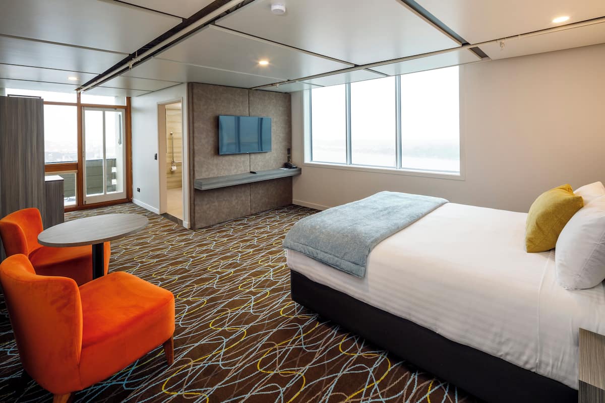 A hotel room with a bed, a TV mounted on the wall, a seating area with a table and chairs, and a large window providing a view.