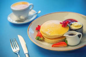 A plate of pancakes with strawberries and a small pitcher of syrup, a cup of coffee, and a fork and knife on a blue table.