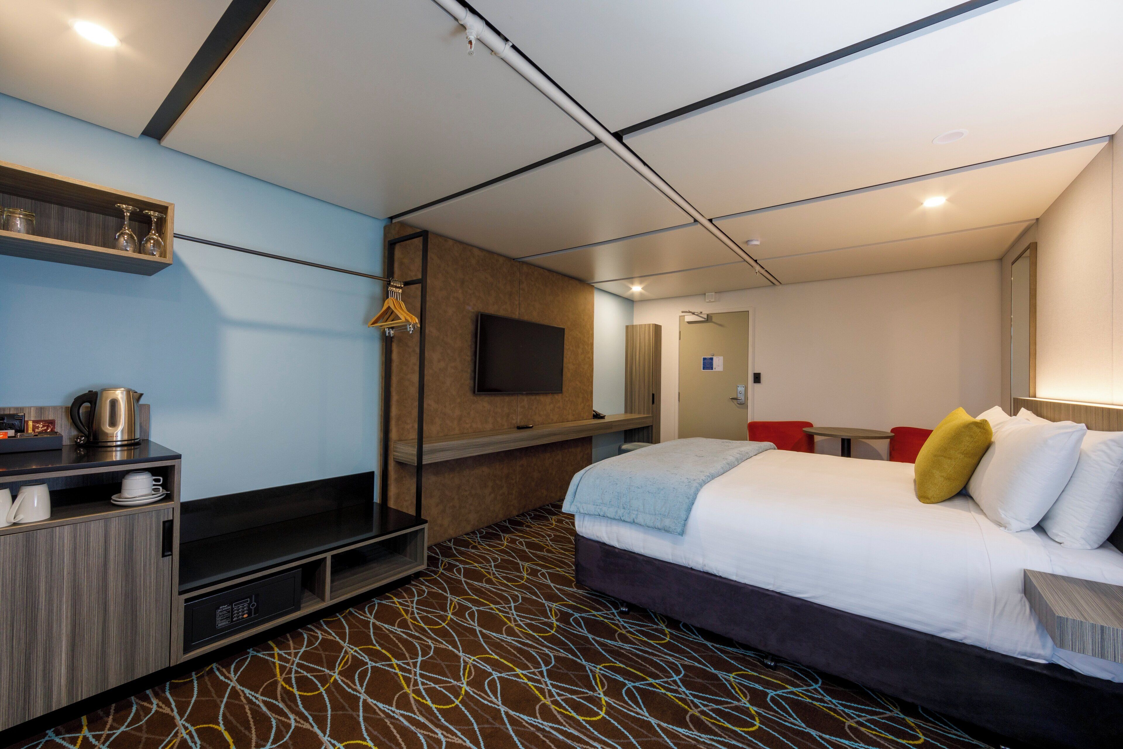 Ein modernes Hotelzimmer mit Bett, Fernseher, Schreibtisch mit Wasserkocher und einer kleinen Sitzecke.