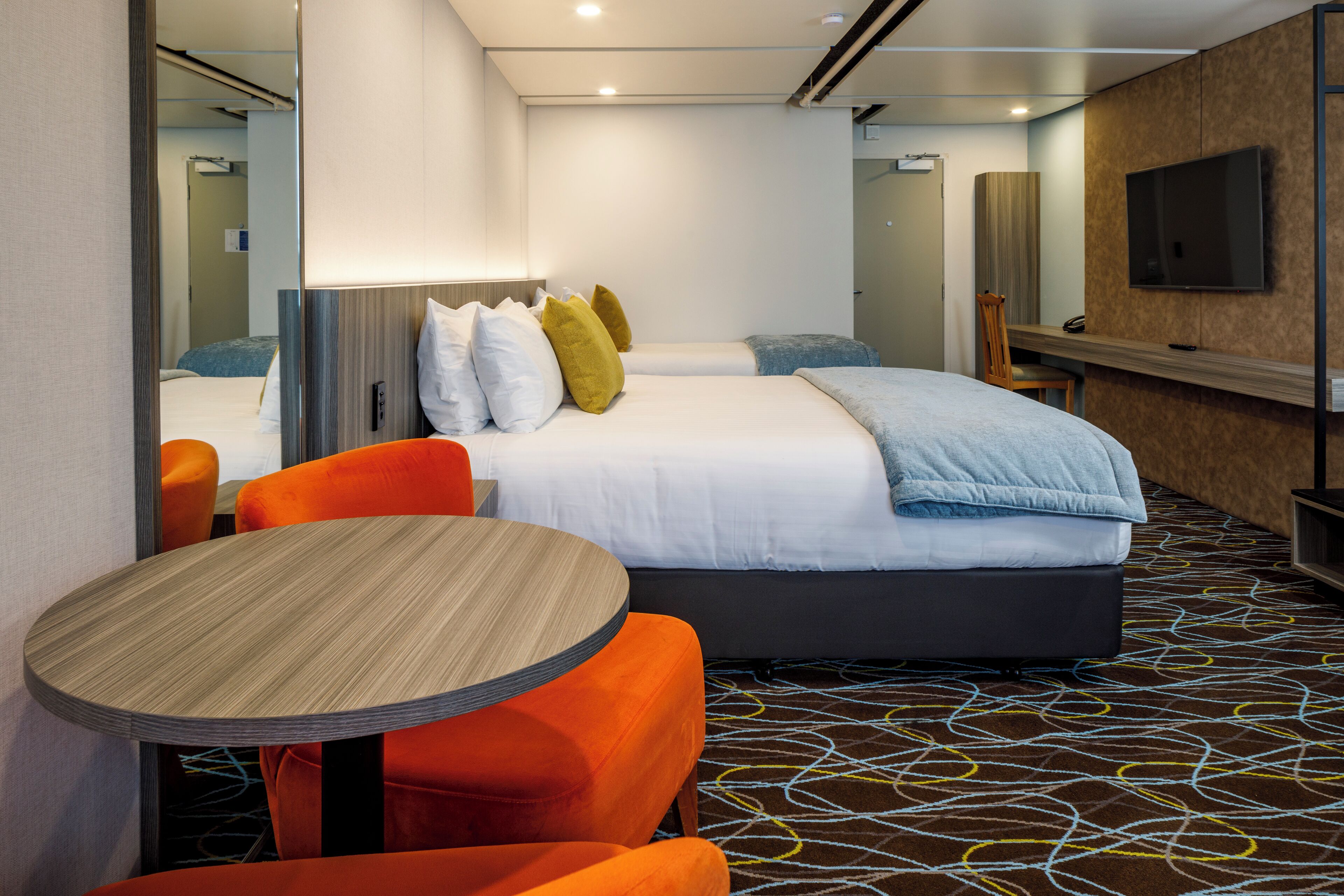 Habitación de hotel con una cama, dos sillones naranjas, una mesa redonda, un televisor y una alfombra estampada.