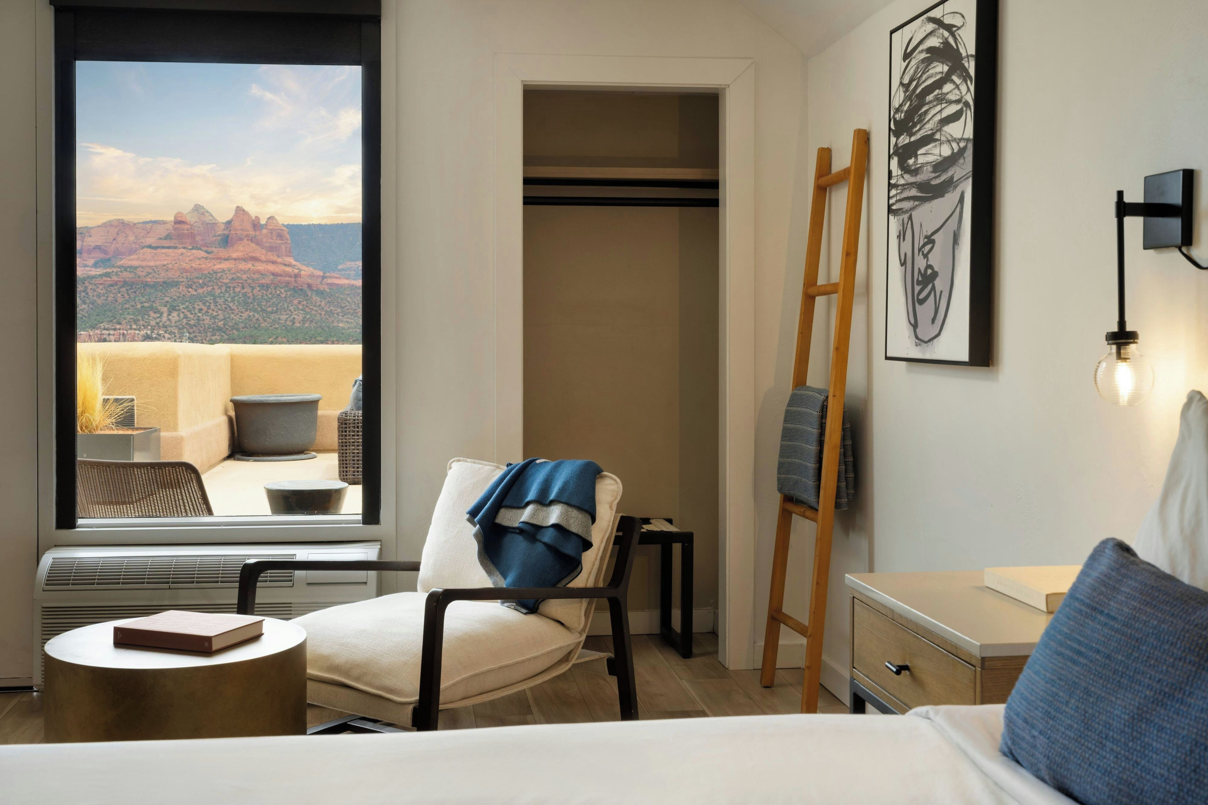 A hotel room with a bed, a chair, a desk, a window with a view of red rock formations, and a ladder leaning against the wall.
