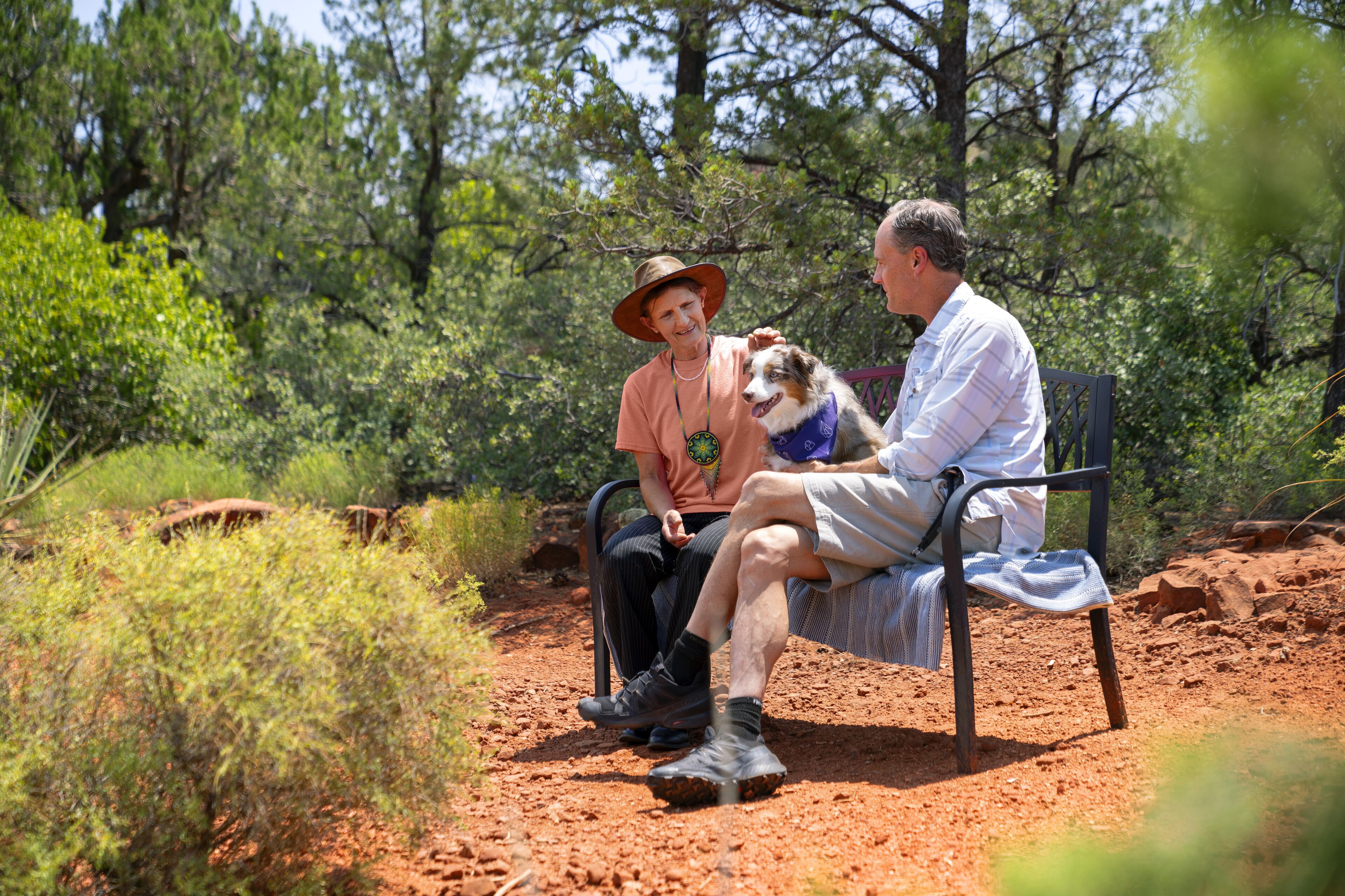 Two people sitting on a bench in a natural setting with trees and shrubs.