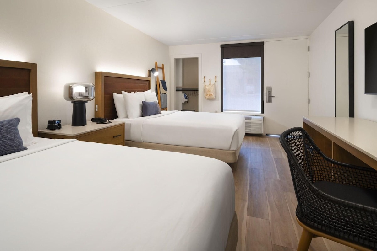 A hotel room with two beds, a desk, a chair, a lamp, a coat rack, and a window.