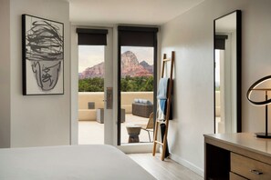 A bedroom with a bed, a desk, a chair, a painting, and a view of mountains through an open door. - Sky Rock Sedona, a Tribute Portfolio Hotel (Sedona)
