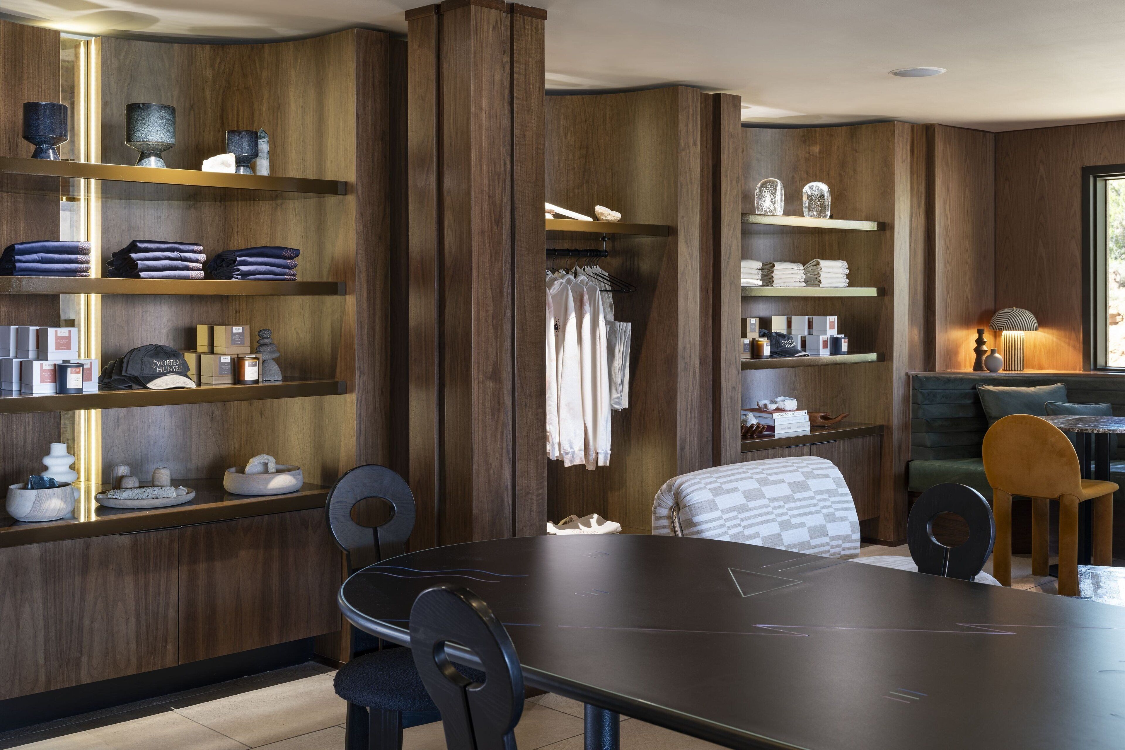 A modern hotel room with wooden paneling, a dining area with a dark table and chairs, and shelves with neatly folded towels and toiletries.