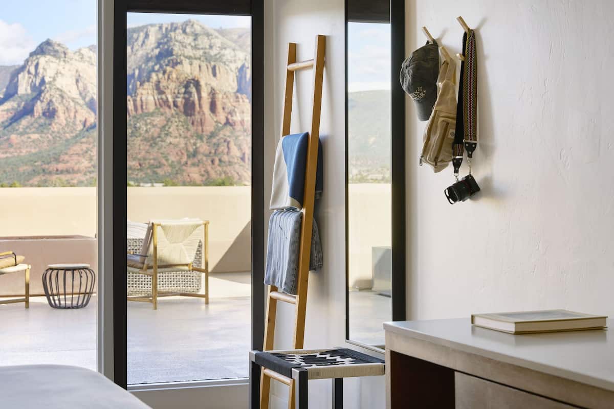 Una habitación con un amplio ventanal que da a un paisaje montañoso, una escalera de madera, una silla y un bolso colgando.