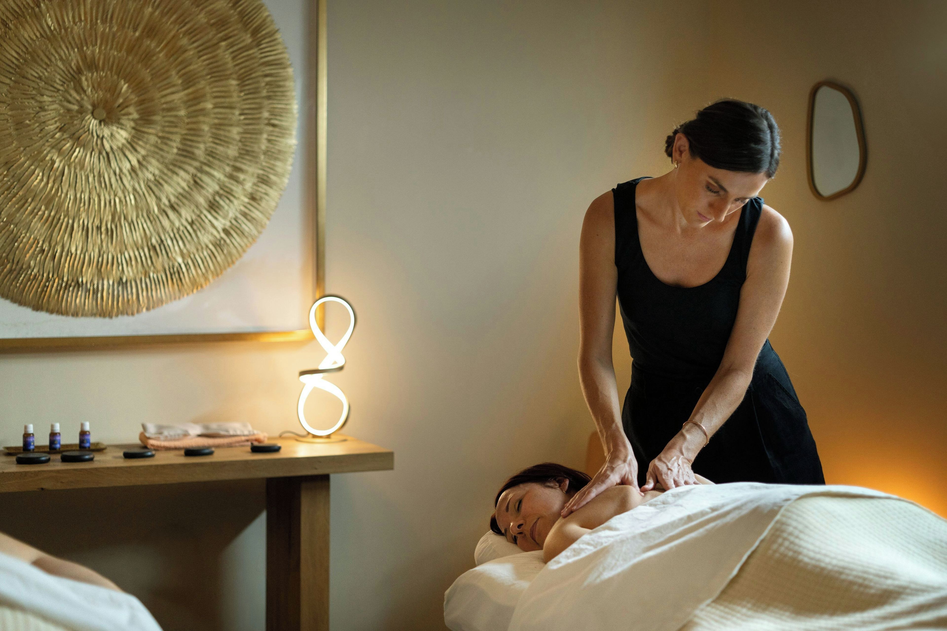 A massage therapy session in a room with a massage table, a small table with bottles, and a wall-mounted light fixture.