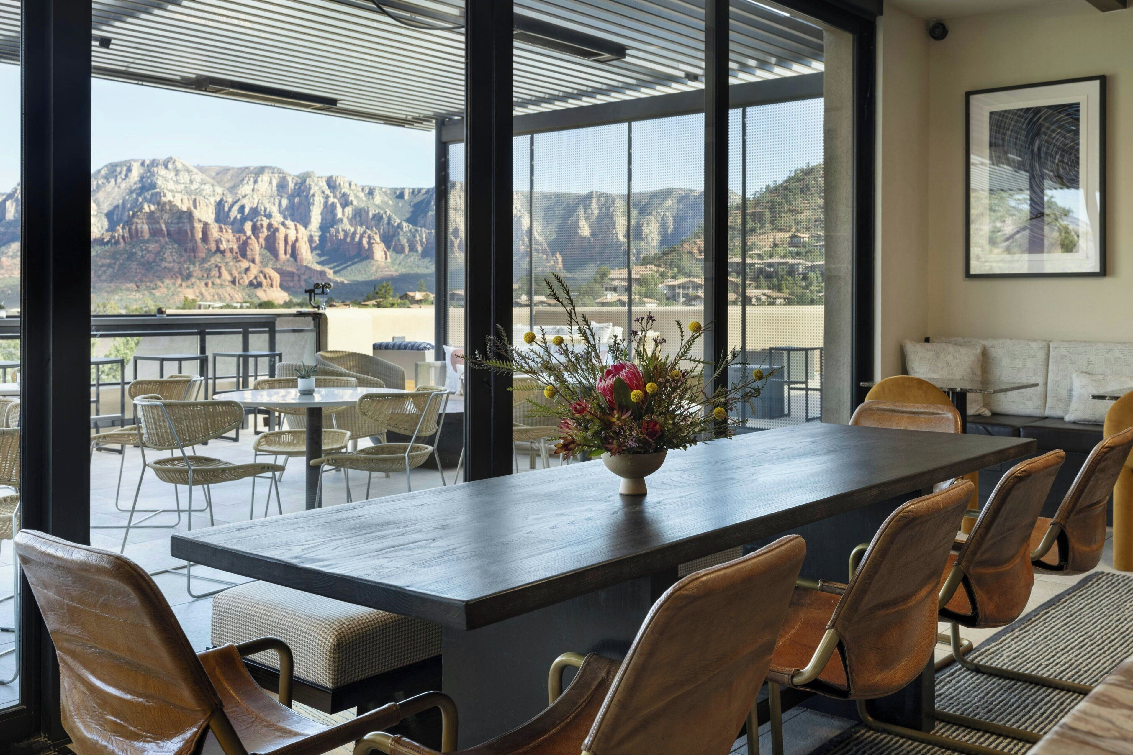 A dining area with a large table and chairs, a flower vase, and a view of mountains through large windows.