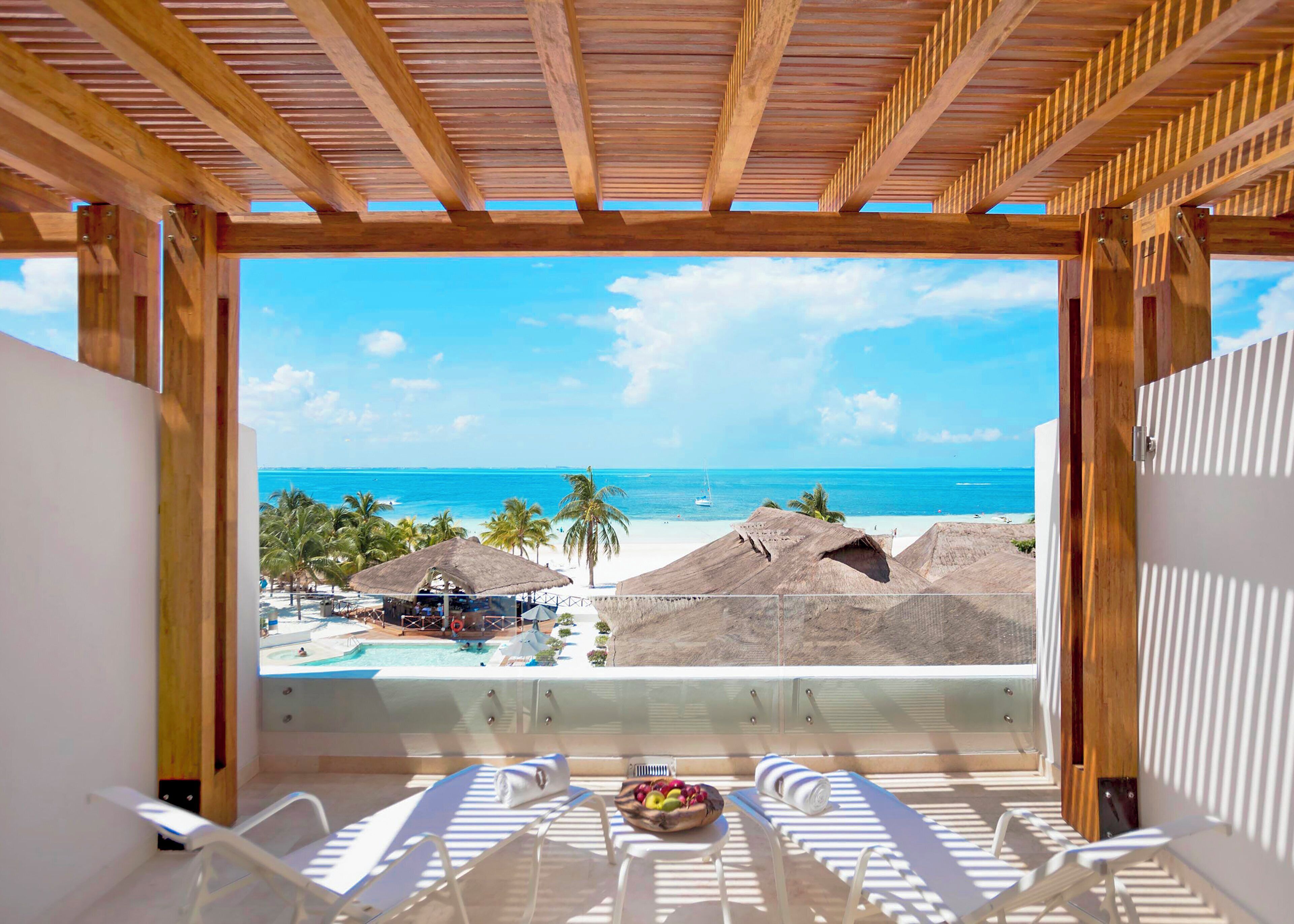 Vista a la playa desde un balcón con vigas de madera, piscina y sillones de descanso.