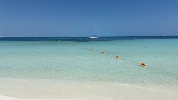 Una playa con agua turquesa cristalina, una orilla arenosa y un velero fondeado en la distancia.