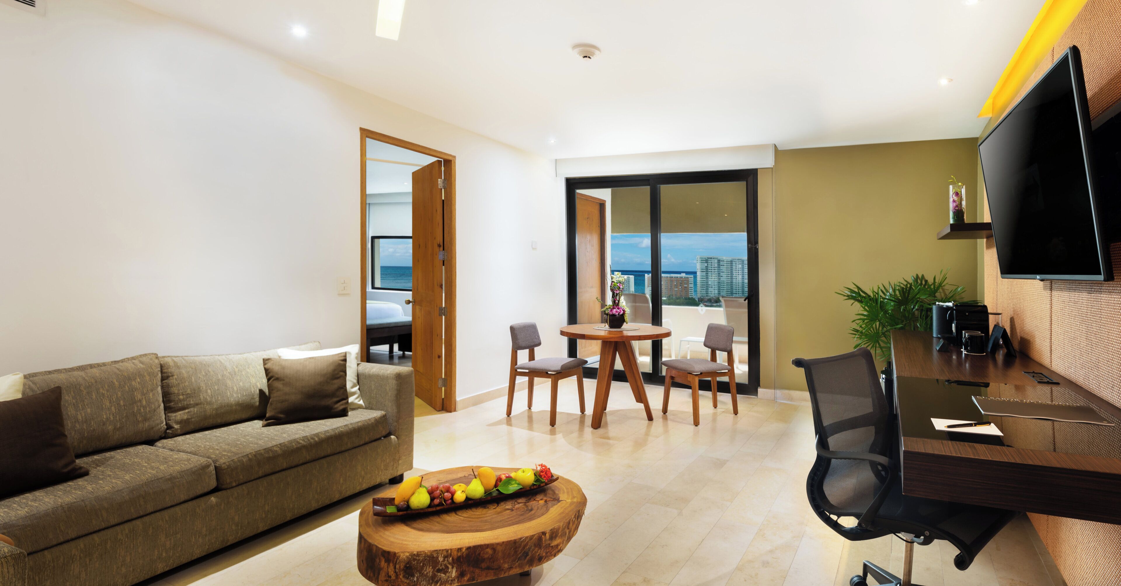 Una sala de estar moderna con un sofá, una mesa de comedor con sillas, una mesa de centro de madera con fruta y un televisor en la pared.