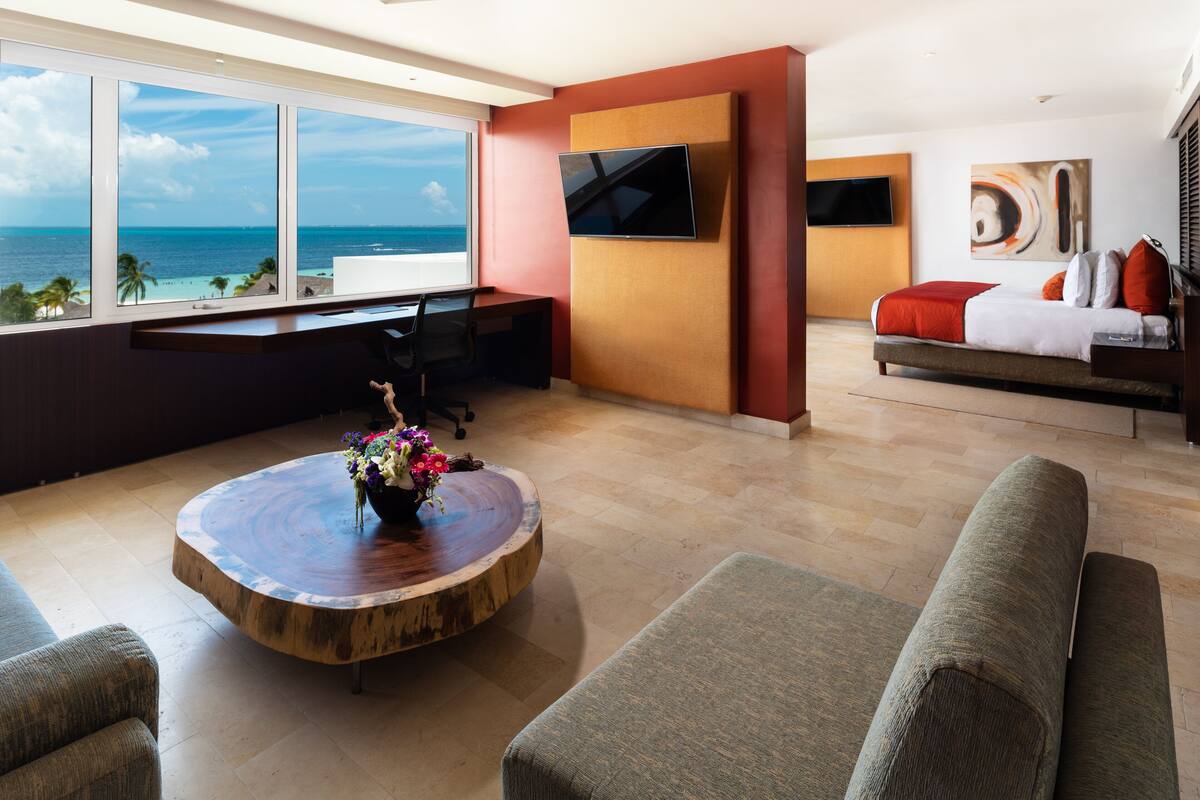 A modern living room with a large window overlooking the ocean, a flat-screen TV mounted on the wall, and a cozy seating area with a round wooden coffee table and colorful flowers.