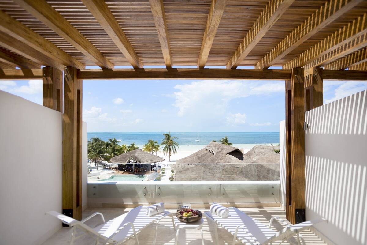 A balcony with a view of the beach and ocean, white lounge chairs, and a table with fruit.