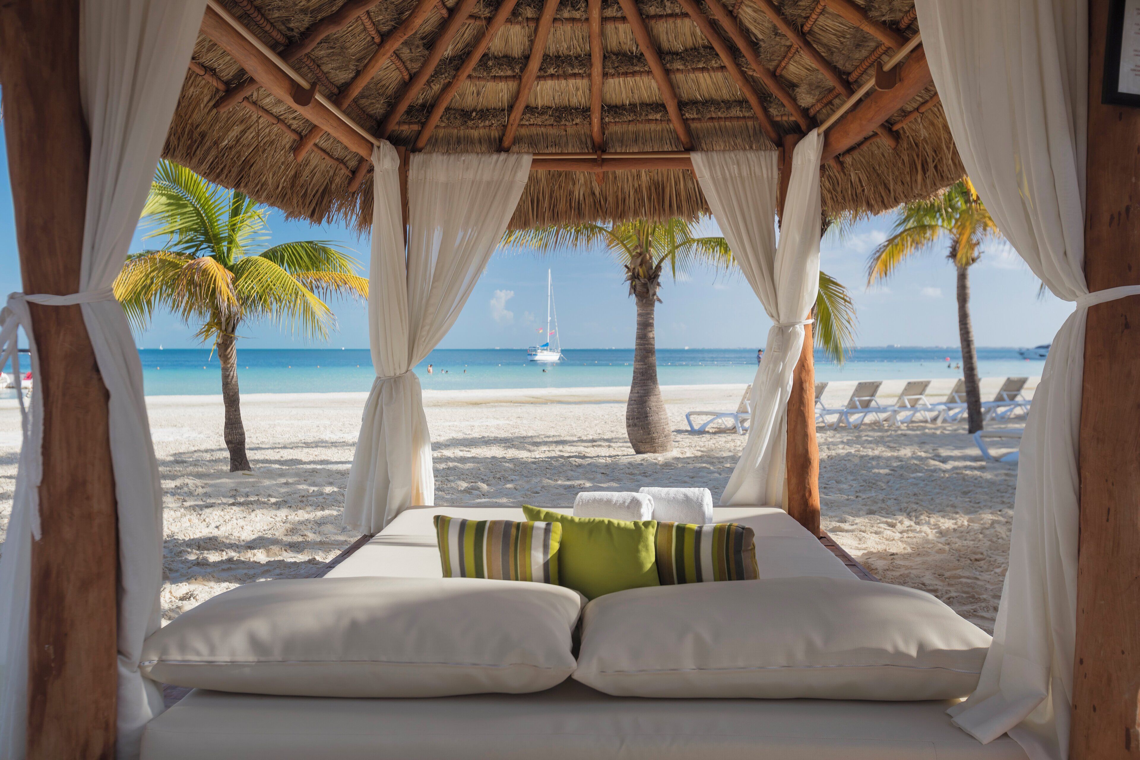 Blick auf einen Strand vom Fenster einer Hütte mit Reetdach, mit Bett, Kissen und Vorhängen.