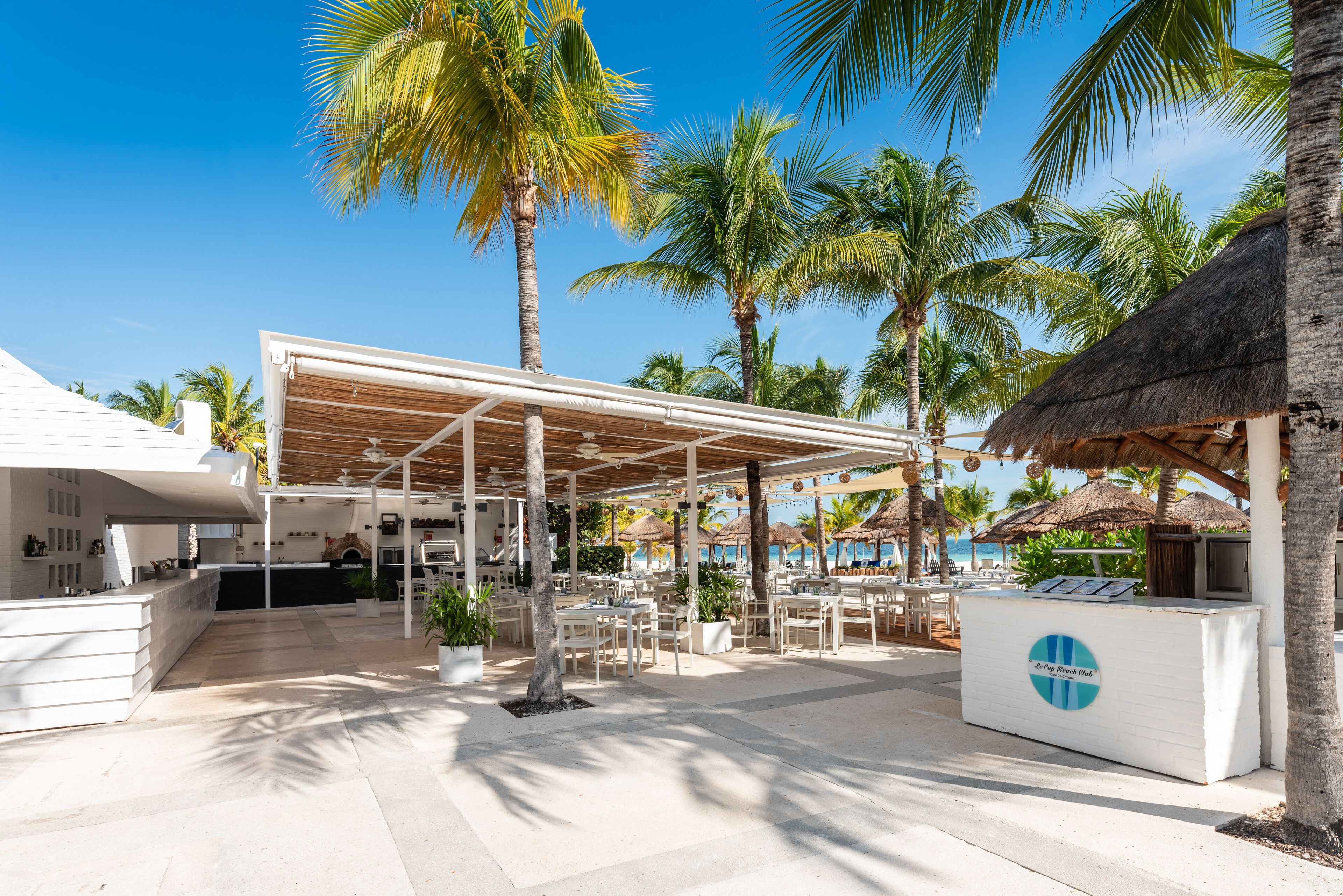 A beachside resort with outdoor seating, palm trees, and a thatched-roof structure.
