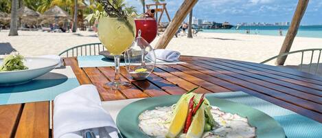 Una mesa de madera con ceviche, una copa de vino blanco y un cóctel en un entorno de playa.