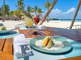 Una mesa de madera con ceviche, una copa de vino blanco y un cóctel en un entorno de playa.