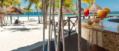 A wooden deck with rope swings, a bar area with a drink, and a beach with palm trees and a thatched roof structure.