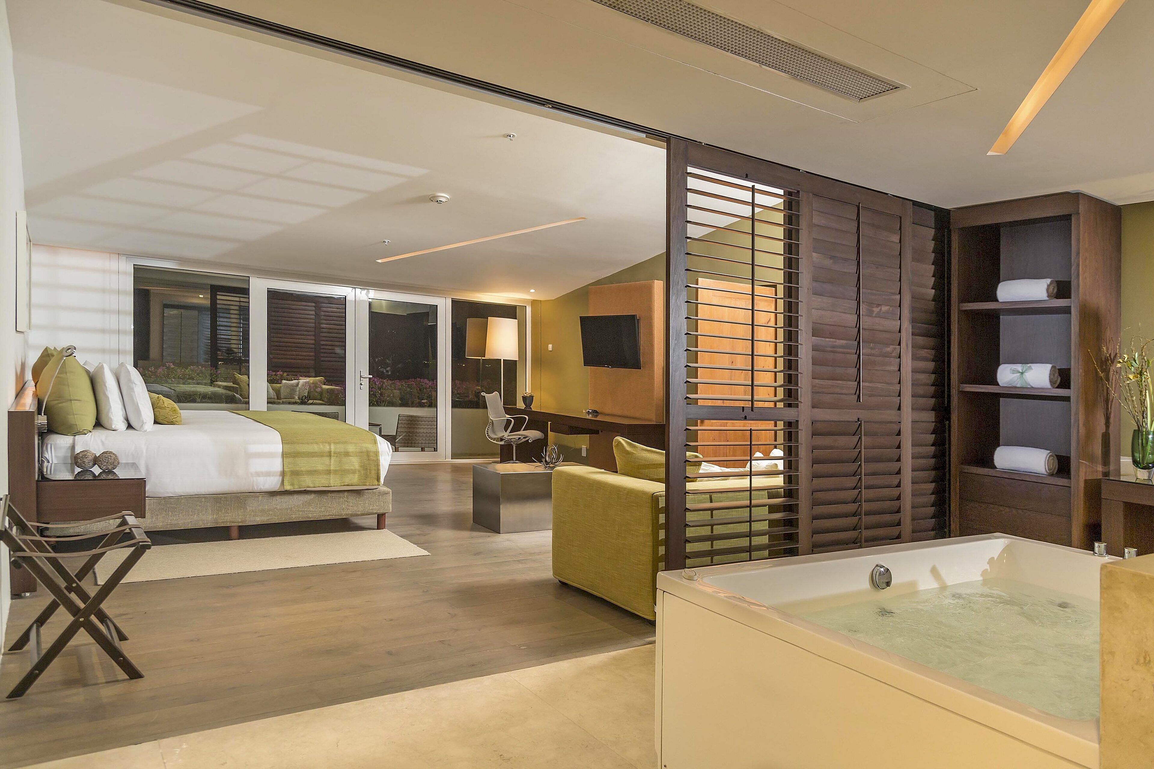 A modern hotel room with a large bed, a sitting area with a sofa and armchairs, a bathtub, and a wooden partition.
