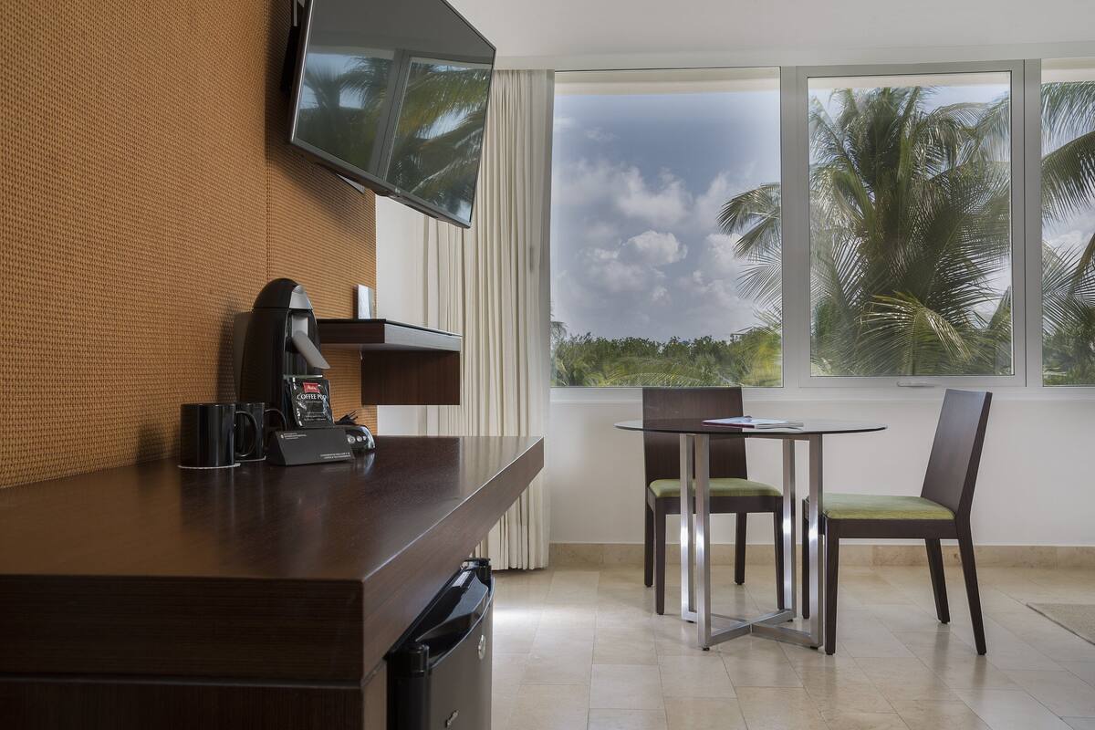 A hotel room with a kitchenette, a dining area with a table and chairs, a large window with a view of palm trees, and a flat-screen TV mounted on the wall.