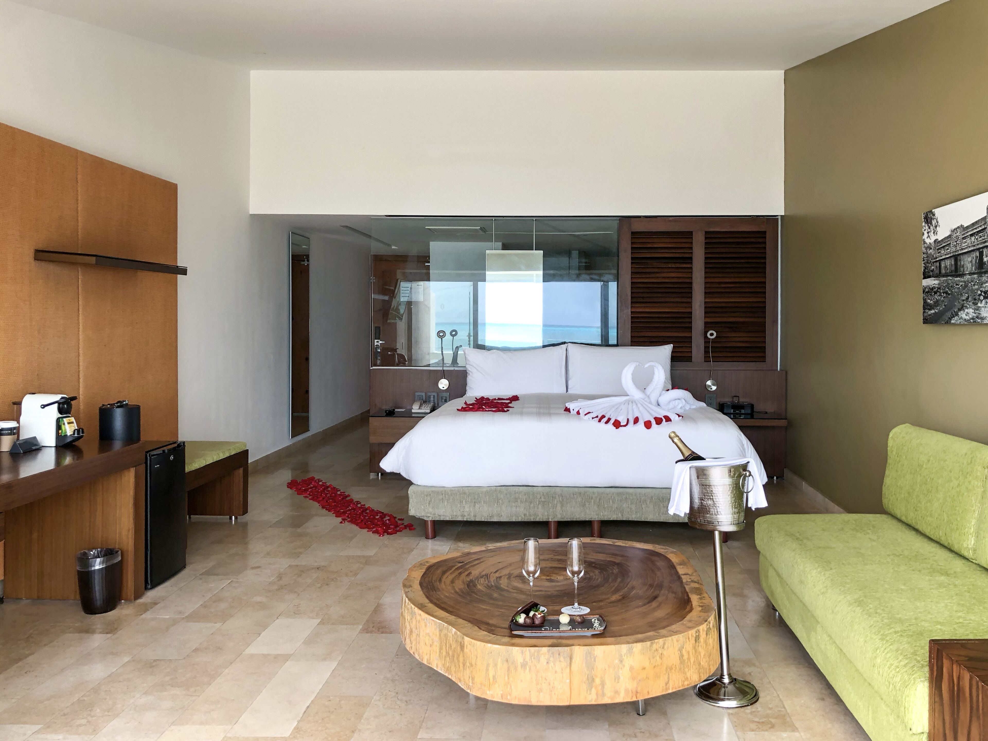 A hotel room with a bed, a green sofa, a wooden coffee table with glasses, a trash bin, and a minibar.