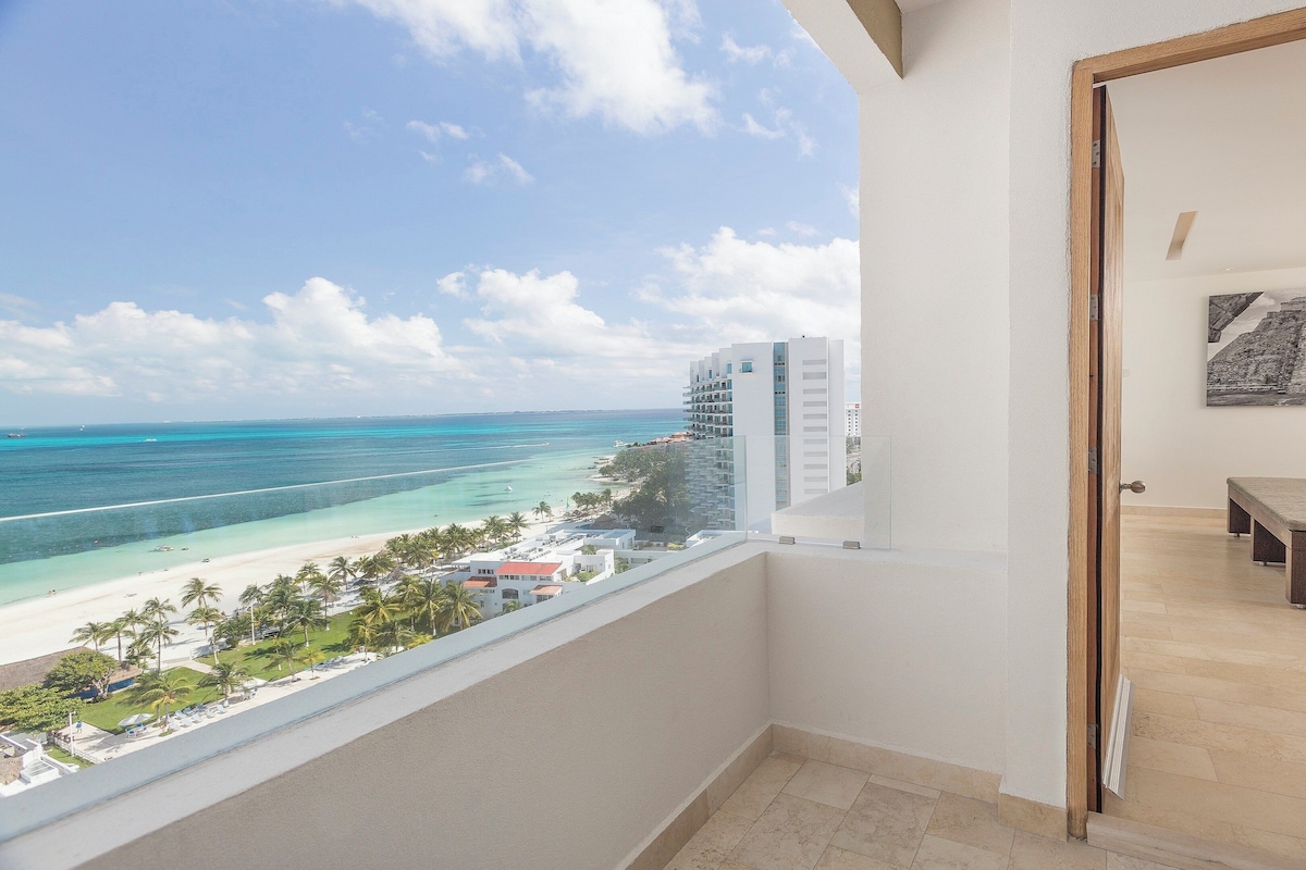 A balcony with a view of a beach and ocean, a bed with a bench, and a painting on the wall.